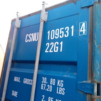A close-up view of a blue shipping container door displaying white text with serial numbers and weight specifications. The metal bolts and latches are visible, adding a rugged aspect to the industrial container.