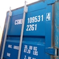 A close-up view of a blue shipping container door displaying white text with serial numbers and weight specifications. The metal bolts and latches are visible, adding a rugged aspect to the industrial container.