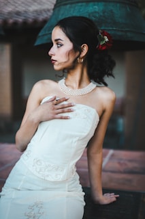 A young woman in a white, embroidered strapless dress poses elegantly with her hand on her chest. Her hair is styled with a red flower accessory, and she has dark eye makeup and red lipstick. The background features a large bell and architectural elements.