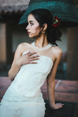 A young woman in a white, embroidered strapless dress poses elegantly with her hand on her chest. Her hair is styled with a red flower accessory, and she has dark eye makeup and red lipstick. The background features a large bell and architectural elements.