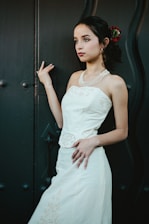 A young woman stands gracefully against a textured dark background, wearing a white dress with intricate embroidery and a pearl necklace. Her hair is styled elegantly with a red flower tucked in.