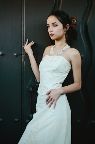 A young woman stands gracefully against a textured dark background, wearing a white dress with intricate embroidery and a pearl necklace. Her hair is styled elegantly with a red flower tucked in.