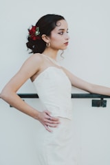 A young woman in a white formal dress stands in profile against a plain light background. Her dark hair is styled with red and green floral accessories, and she wears gold hoop earrings and a subtle necklace. Her expression is calm and contemplative, with her hands resting on a railing.