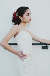 A young woman in a white formal dress stands in profile against a plain light background. Her dark hair is styled with red and green floral accessories, and she wears gold hoop earrings and a subtle necklace. Her expression is calm and contemplative, with her hands resting on a railing.
