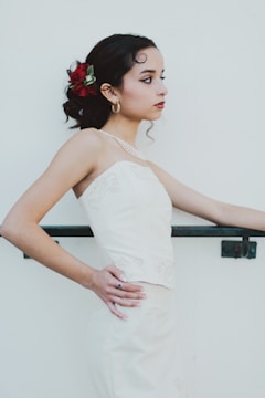 A young woman in a white formal dress stands in profile against a plain light background. Her dark hair is styled with red and green floral accessories, and she wears gold hoop earrings and a subtle necklace. Her expression is calm and contemplative, with her hands resting on a railing.
