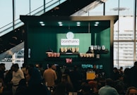 A bustling cafe scene with a modern design, featuring a counter adorned with various coffee products and a logo reading 'bonmano' under well-placed lights. Shelves display neatly arranged packages of goods, while a diverse crowd of people engage in lively conversations or queue up.