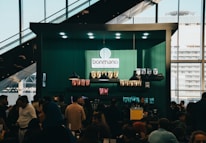 A bustling cafe scene with a modern design, featuring a counter adorned with various coffee products and a logo reading 'bonmano' under well-placed lights. Shelves display neatly arranged packages of goods, while a diverse crowd of people engage in lively conversations or queue up.