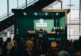 A bustling cafe scene with a modern design, featuring a counter adorned with various coffee products and a logo reading 'bonmano' under well-placed lights. Shelves display neatly arranged packages of goods, while a diverse crowd of people engage in lively conversations or queue up.