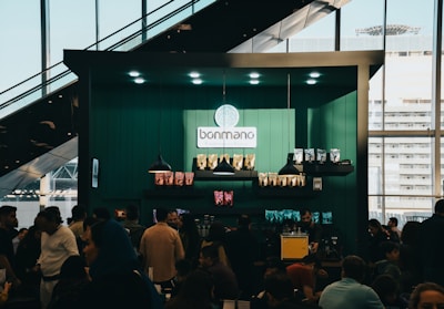 A bustling cafe scene with a modern design, featuring a counter adorned with various coffee products and a logo reading 'bonmano' under well-placed lights. Shelves display neatly arranged packages of goods, while a diverse crowd of people engage in lively conversations or queue up.
