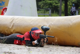 A person wearing protective gear, including a red helmet and jersey, is lying on the ground aiming a paintball gun. They are positioned behind a large inflatable barrier on a paintball field, which has some greenery in the background.