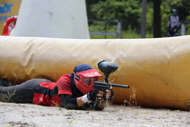 A person wearing protective gear, including a red helmet and jersey, is lying on the ground aiming a paintball gun. They are positioned behind a large inflatable barrier on a paintball field, which has some greenery in the background.