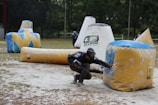 A paintball player crouches behind a large inflatable obstacle, wearing protective gear and holding a paintball marker. The setting is an outdoor field scattered with various inflatable bunkers. Another player is visible in the background, also taking cover.