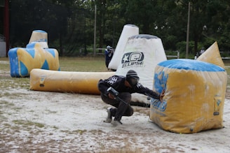 A paintball player crouches behind a large inflatable obstacle, wearing protective gear and holding a paintball marker. The setting is an outdoor field scattered with various inflatable bunkers. Another player is visible in the background, also taking cover.