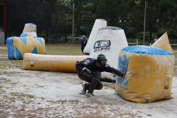 A paintball player crouches behind a large inflatable obstacle, wearing protective gear and holding a paintball marker. The setting is an outdoor field scattered with various inflatable bunkers. Another player is visible in the background, also taking cover.