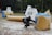 A paintball player crouches behind a large inflatable obstacle, wearing protective gear and holding a paintball marker. The setting is an outdoor field scattered with various inflatable bunkers. Another player is visible in the background, also taking cover.