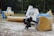 A paintball player crouches behind a large inflatable obstacle, wearing protective gear and holding a paintball marker. The setting is an outdoor field scattered with various inflatable bunkers. Another player is visible in the background, also taking cover.