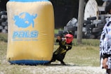 A person wearing a red mask and a black and yellow outfit is crouched behind a large inflatable barrier with the text 'PALS Paintball-Asia League Series' on it. The person is holding a paintball gun, aiming forward. The field includes grass and scattered tires, suggesting a paintball arena.
