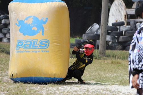 A person wearing a red mask and a black and yellow outfit is crouched behind a large inflatable barrier with the text 'PALS Paintball-Asia League Series' on it. The person is holding a paintball gun, aiming forward. The field includes grass and scattered tires, suggesting a paintball arena.