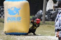 A person wearing a red mask and a black and yellow outfit is crouched behind a large inflatable barrier with the text 'PALS Paintball-Asia League Series' on it. The person is holding a paintball gun, aiming forward. The field includes grass and scattered tires, suggesting a paintball arena.