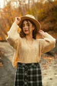 A woman stands outdoors wearing a knitted beige sweater and a black and white plaid skirt, with one hand adjusting her wide-brimmed brown hat. Her makeup is bold with red lipstick and brightly colored eyeshadow. The background features a blurred autumnal landscape with trees full of orange and yellow leaves.