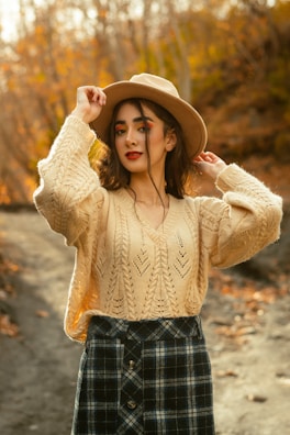 A woman stands outdoors wearing a knitted beige sweater and a black and white plaid skirt, with one hand adjusting her wide-brimmed brown hat. Her makeup is bold with red lipstick and brightly colored eyeshadow. The background features a blurred autumnal landscape with trees full of orange and yellow leaves.