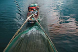 A wooden boat laden with various supplies and equipment is navigating through calm waters, pulling a large fishing net. The surface of the water reflects shades of blue and pink.