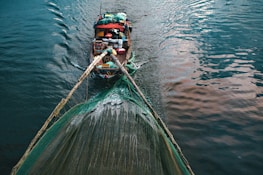 A wooden boat laden with various supplies and equipment is navigating through calm waters, pulling a large fishing net. The surface of the water reflects shades of blue and pink.