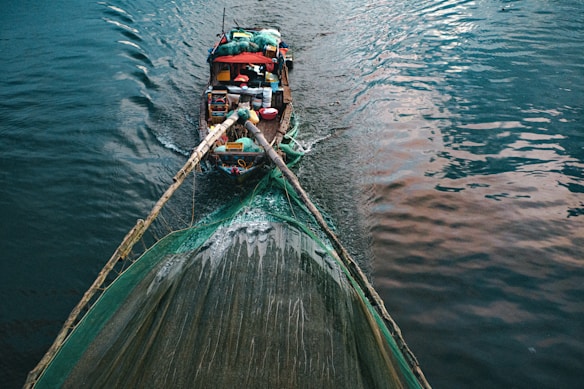 A wooden boat laden with various supplies and equipment is navigating through calm waters, pulling a large fishing net. The surface of the water reflects shades of blue and pink.