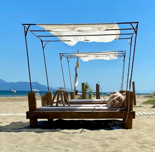 A serene beach scene featuring a wooden lounge structure draped with white fabric. Sandy beach stretches towards calm blue waters, with mountains visible on the horizon. A couple of small boats float in the sea, and a few decorative pillows are placed on the lounge bed.