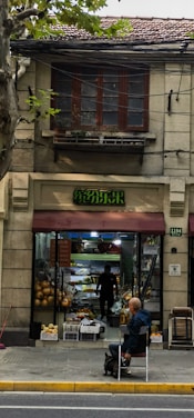 A fruit shop with an open entrance, showcasing various fruits like pumpkins displayed outside. A person inside is arranging produce, and outside, a bald man is sitting on a chair with a small dog on a leash beside him. The building features a rustic facade with an overhanging awning in a muted red tone.
