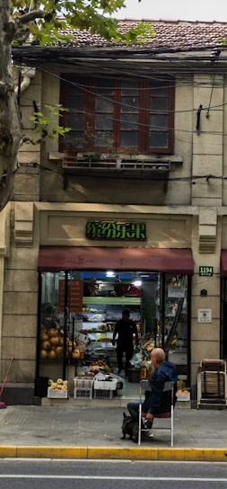 A fruit shop with an open entrance, showcasing various fruits like pumpkins displayed outside. A person inside is arranging produce, and outside, a bald man is sitting on a chair with a small dog on a leash beside him. The building features a rustic facade with an overhanging awning in a muted red tone.