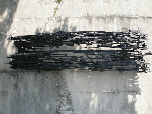 A large assortment of black rifles is neatly arranged in parallel rows on a concrete surface. The image is taken from above, casting shadows on the rough texture of the ground. The firearms vary in size and are tightly packed together, indicating a focus on military or weapon-related themes.