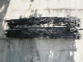 A large assortment of black rifles is neatly arranged in parallel rows on a concrete surface. The image is taken from above, casting shadows on the rough texture of the ground. The firearms vary in size and are tightly packed together, indicating a focus on military or weapon-related themes.