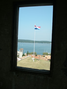 A flagpole with the Croatian flag is prominently visible against a clear blue sky. The scene is framed by a rectangular window, showing a landscape with a church tower, rooftops, some greenery, and a view of a calm body of water.