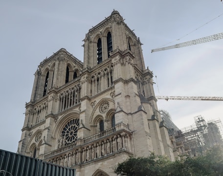 A Gothic-style cathedral with two prominent towers and intricate architectural details, surrounded by construction cranes and scaffolding, indicating restoration work.
