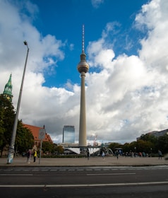 A tall television tower stands prominently against a backdrop of a partly cloudy blue sky. Surrounding the tower are buildings and a plaza with people walking and standing around. A church tower is visible to the left, and there are trees and city infrastructure such as a road and streetlights.