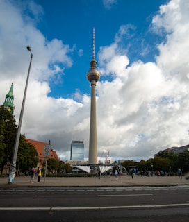 A tall television tower stands prominently against a backdrop of a partly cloudy blue sky. Surrounding the tower are buildings and a plaza with people walking and standing around. A church tower is visible to the left, and there are trees and city infrastructure such as a road and streetlights.