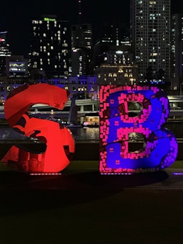 Large illuminated letters 'S' and 'B' are prominently displayed in the foreground, with vibrant red and blue lighting. The background features a cityscape with tall buildings, some of which are lit with colorful lights, against a night sky.