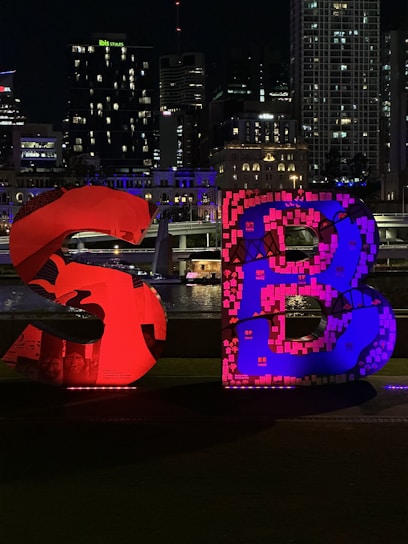 Large illuminated letters 'S' and 'B' are prominently displayed in the foreground, with vibrant red and blue lighting. The background features a cityscape with tall buildings, some of which are lit with colorful lights, against a night sky.
