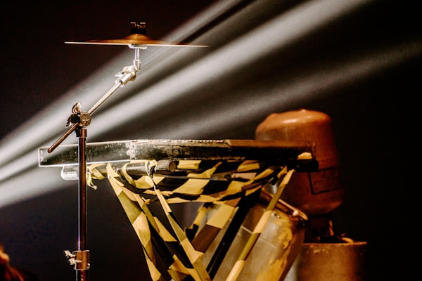 A cymbal mounted on a stand is illuminated by stage lighting, casting sharp beams across the scene. The setup features a table reinforced with yellow and black caution tape, possibly indicating an industrial or makeshift theme. An object resembling a gas canister is seen nearby, contributing to the mechanical aesthetic.