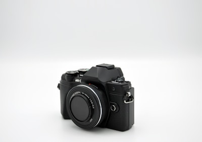 A black digital camera with a large lens is positioned on a plain white background. The camera features manual controls and a textured grip, suggesting it is designed for advanced photography.
