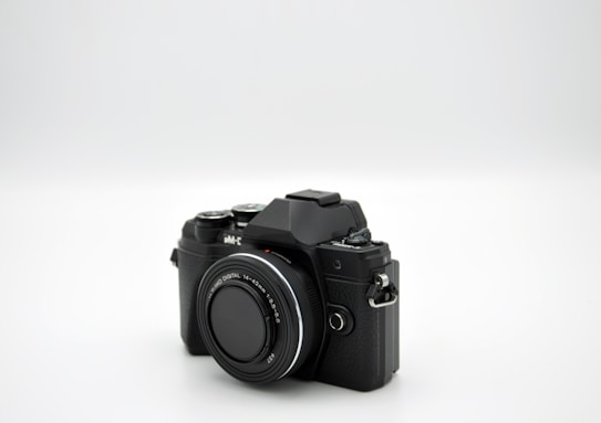 A black digital camera with a large lens is positioned on a plain white background. The camera features manual controls and a textured grip, suggesting it is designed for advanced photography.