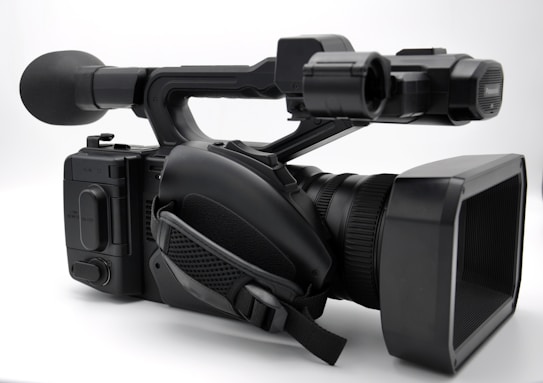 A professional video camera with a large lens and a microphone attached is positioned on a white background. The camera has a robust design with various controls and a carrying handle.