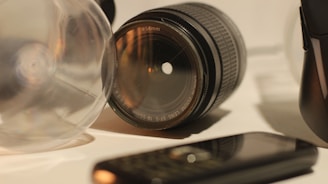 A close-up of a camera lens is surrounded by various objects, including a transparent glass-like object and a mobile phone lying on its front. The focus is on the lens, highlighting its details and reflections in a warm, subdued light.