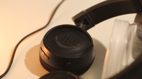 A close-up view of black over-ear headphones with a visible JBL logo on the ear cup, positioned on a light surface. The headphones are partially coiled with a cable stretching away from the headset.