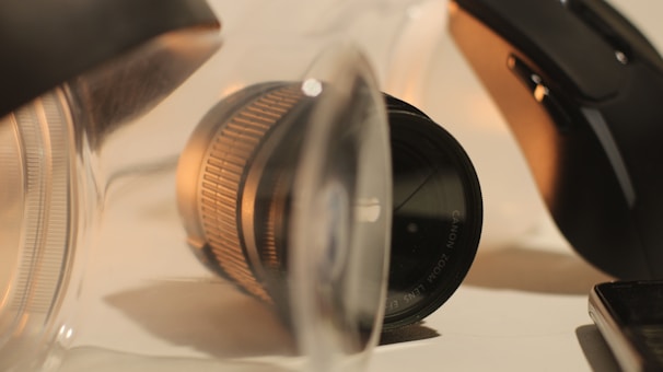 A close-up view of a DSLR camera lens partially enclosed in a transparent plastic container, with a computer mouse and part of a smartphone also visible. The lighting casts warm tones on the objects, and the focus is on the texture and detail of the lens.