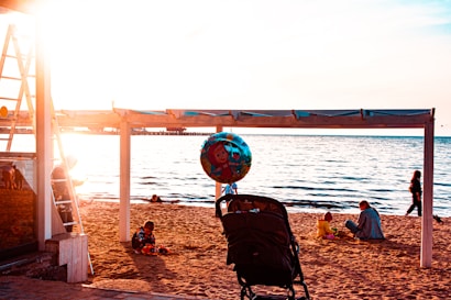 A sunlit beach scene features a child playing with toy cars on the sand. A colorful balloon is attached to a stroller in the foreground. Several people are sitting and walking along the beach, enjoying the warm light of the setting sun. In the background, the calm sea and a distant pier are visible.