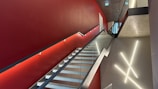 A modern interior staircase with glass railings and a red accent wall. The wall features minimalist lighting fixtures arranged in a geometric pattern. The ceiling is concrete with recessed lighting, and a doorway is visible at the top of the stairs.