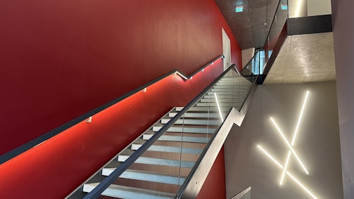 A modern interior staircase with glass railings and a red accent wall. The wall features minimalist lighting fixtures arranged in a geometric pattern. The ceiling is concrete with recessed lighting, and a doorway is visible at the top of the stairs.