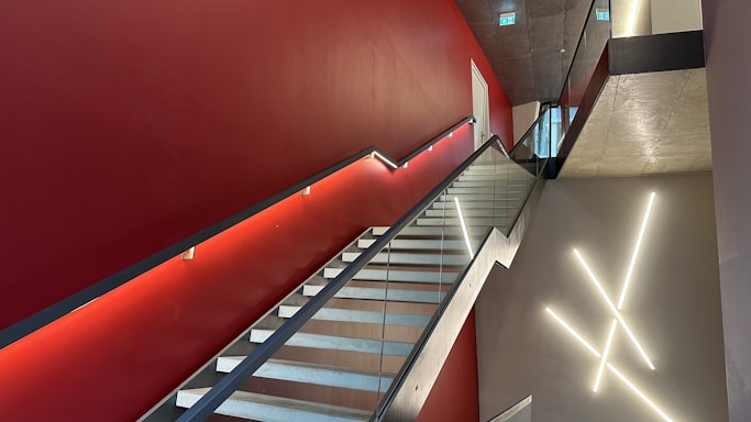A modern interior staircase with glass railings and a red accent wall. The wall features minimalist lighting fixtures arranged in a geometric pattern. The ceiling is concrete with recessed lighting, and a doorway is visible at the top of the stairs.